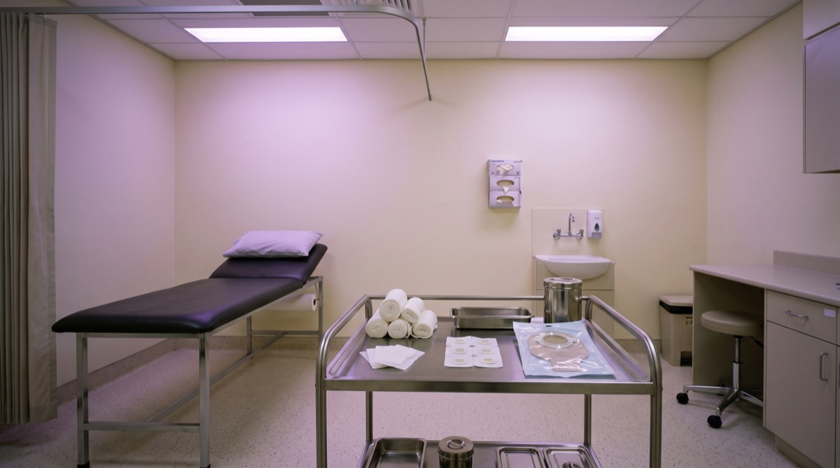 An empty hospital examination room lit with soft violet ambient light from recessed ceiling panels, a stainless steel supply trolley holding rolled gauze, adhesive flange squares, and a sealed stoma pouch in a neat clinical arrangement, pale cream walls, an empty padded examination table to one side, no people, no text visible on any surface, depopulated clinical setting, photographic realism