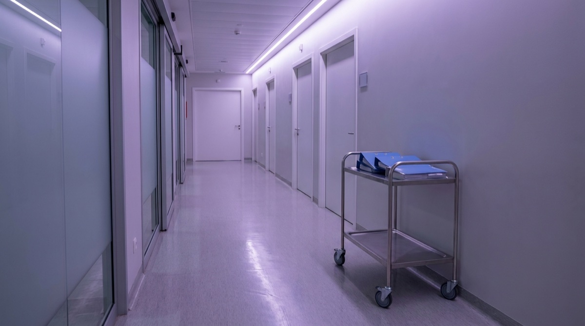 A quiet clinical research corridor inside a modern hospital, soft violet ambient light filtering through frosted glass partitions, a row of closed white treatment room doors, a stainless steel trolley with sealed research binders placed neatly against the wall, clean linoleum floor reflecting the overhead lights, no people visible, no readable text on any surface, depopulated medical setting, photographic